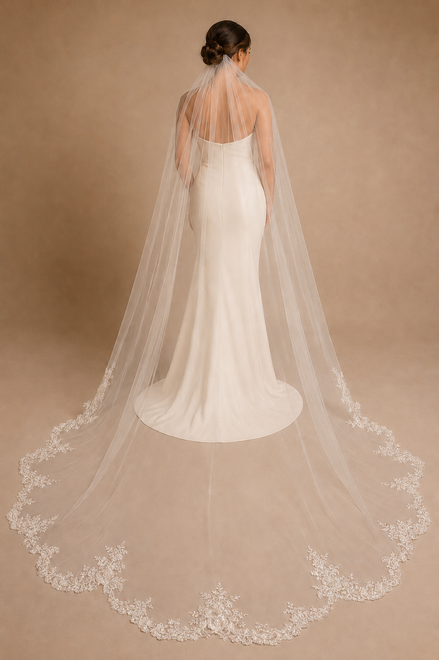 A bride viewed from behind wearing a white wedding gown and a long sheer tulle bridal veil with floral lace trim, standing against a warm beige studio backdrop.